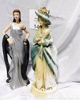 Two female statuettes shown side by side, one modern and one vintage, approximately 10 inches tall each, against a white cloth background.