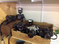 Two cameras (Pentax and Minolta) on shelf above cardboard box containing multiple cameras and a flash unit