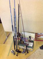 Photo showing four fishing rods with different handle colors leaning against a wall, three fishing reels on the floor, and a green tackle box filled with fishing gear.