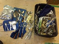 Overview of assorted silver flatware laid out on floor including forks, spoons, handles of darker colored material, and a bag with additional pieces.