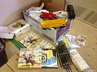 Box containing French Canadian trivia games with colorful traditional images, model trains including yellow boxcar and black train part, model train rails, and remote control devices.