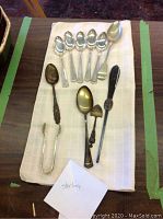 Full view of all spoons and silver tongs laid out on white cloth