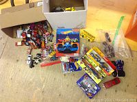 Wide view of various small diecast automotive toys scattered on the floor with two open boxes including boxed Hot Wheels cars and loose pieces.