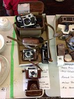 Three vintage 35mm cameras (Voigtlander Vito Automatic II, Tenax Pronormat S, Yashica YK 1031052) with leather cases and accessories on table.