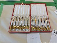 Two open red boxes containing six gold-tone enamel spoons each, with hand-painted decoration and satin lining.