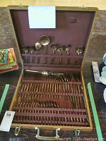 Open wooden case showing brass cutlery including spoons, forks, and ladles laid in fitted slots.