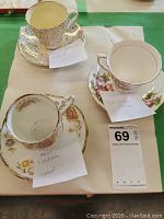 Three different teacup and saucer sets on a white mat with small note labels identifying makers and signatures