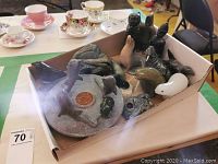 Cardboard box containing multiple soapstone figurines including seals, walrus, and other marine animals, placed on a green surface with tea cups in the background.