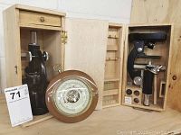 Two vintage black metal microscopes in separate light wooden cases. Cases have compartments and drawers for accessories and glass slides. A round wooden-framed barometer is placed in front of one case.