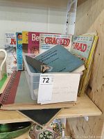 View of assorted vintage magazines and postcards in a container on a wooden shelf. Visible titles include Mad magazine, Cracked, Beverly, and Super magazines, along with a blue booklet and spiral notebook.