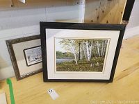 Two framed prints and one framed painting together on floor against wall, one print smaller and one larger painting with birch tree scene