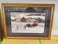 Framed Walter Campbell print showing a winter scene of a horse-drawn sleigh carrying people past a red house in snowy countryside.