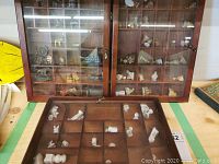 Photo showing a wooden case with multiple compartments holding white ceramic or porcelain miniature figures, some damaged.