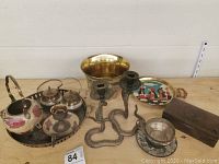 Photo showing multiple brass and enamel items including small kettles, a planter, snake-shaped candle holders, a small brass cup, and a decorative box on a wooden surface.