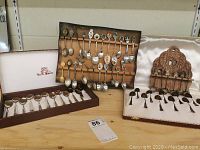 Photo showing multiple collector's spoons arranged in groups: vintage spoons in an ornate case on the right, souvenir spoons on a wooden rack center back, and La S Marco spoon set in original box front left.