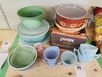 Photo of assorted vintage Pyrex mixing bowls, cups, pink creamer, small bowl, and red-orange Pyrex casserole dish with lid on wicker holder on a wooden shelf.