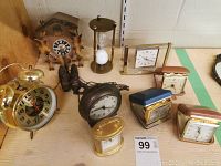 Multiple vintage clocks, including alarm clocks, cuckoo clock with pinecone weights, hourglass timer, and various travel clocks arranged on a shelf
