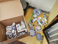 Overview of the box filled with various Delft Blue hand-painted ceramics plus items arranged beside the box on the floor showing a collection of plates, shoe figurines, creamers, mugs, tiles and a small box.