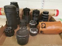 Overall view of assorted lenses with cylindrical cases and lens test chart tube in background