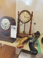 Photo showing two clocks and two replica guns on a shelf. One clock is a mantel style dark wood clock, the other is a taller wooden electric clock with pendulum. Two replica pistols are laid next to clocks.