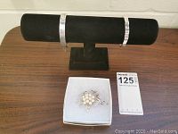 Two narrow silver bracelets displayed on black velvet stand with silver plated brooch in white box on wooden surface.