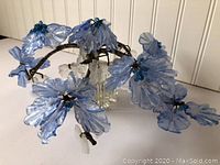 Angled photo showing the blue glass bonsai flowers, clear glass leaves, brown branches, and the square clear glass vase containing the plant.