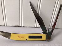 Photo showing Vintage Ka-Bar fish knife with two blades open, yellow handle with metal bolsters and brand name on it.