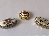 Three vintage decorative pillboxes displayed on a white surface, showing top and side views with floral painted and enameled designs.