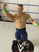 John Cena action figure close-up showing muscular body, green wristbands, and dark blue shorts standing on a WWE circular base