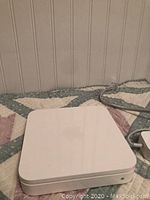 White Apple AirPort Extreme router with rounded edges placed on a quilted fabric surface. Power cord connected, showing the main device.