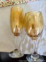 Two clear glass wine and champagne glasses with gold-tone decorative scrollwork patterns on the bowl and a gold accent on the stem, photographed on a white cloth.