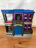 Front and side view of the Batman Gotham City Jail playset structure showing blue double door, balconies, ladder, and windowed jail cells in purple and gray plastic.