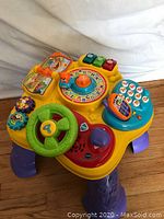 Front angled view of VTech baby play table showing steering wheel, spinning gears, colorful buttons, phone pad, and interactive dial with animal cards, all detailed on the yellow surface with purple legs.