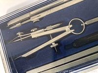 Close-up photo of drafting tools including a large compass with adjustable screw, dividers, and pen holders placed against a blue foam background inside a case.