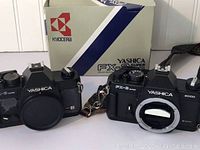 Photo of two Yashica 35mm film camera bodies, one with Yashica FX-3 Super 2000 box, both black cameras shown from front without lenses mounted