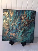 Front close-up of square canvas abstract pour painting with metallic gold and blue-green swirling colors displayed on an easel.