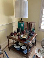 Overall view of lamp with white shade and items arranged on wooden table with lower shelves containing bowls and dishes.