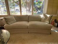 Front view of curved beige fabric sofa with two matching pillows on windowed background showing overall condition and wear.