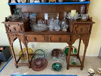 Overview of glassware on antique wooden display table showing bowls, candle holders, plates with some colored glass items on lower shelf.