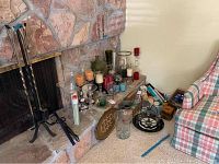 Wide view showing candles, candle holders, vases, trays, and fireplace tools arranged on and near hearth and floor next to a plaid armchair.