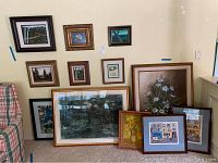 Wide view showing 12 framed art pieces of varying sizes leaning against a wall near a chair. Includes a large floral painting, several smaller landscapes and town scenes with diverse framing styles.
