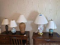 Five assorted table lamps displayed on wood furniture against beige wall, showing variety of bases and shades