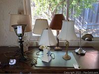 Seven varied table top lamps on wooden desk near window, showing shade and base styles.