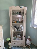 Front view of white wooden display cabinet with contents visible inside and a drawer at the bottom. Shows marks and scratches on surface.