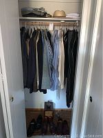 Closet hanging rack with multiple men's tops including jackets and shirts in sizes L to XL, black trousers, and dress shoes on the floor below with a wooden shoe shine box.