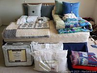 Full lot view with quilts, pillows, white down comforter and pillows in packaging, stacked bedding items