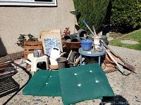 Overall view of wheelbarrow, pots, table, stools, watering can, and lounge cushion outside against wall