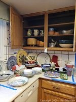 Wide view of kitchen counter and open cabinet showing pie plates, bowls, pitchers, and dishes arranged on countertop and shelves.
