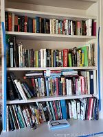 Books arranged on four shelves showing a variety of titles in different genres with some books stacked horizontally and others vertically