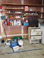 Photo showing small wooden dresser with four drawers, black toolbox, bottles of cleaning and laundry products, jars of nuts and bolts, mop and 5-gallon buckets on the floor